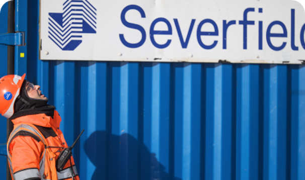 Employee in hardhat and work clothing looking up with blue Severfield branded shipping container in the background
